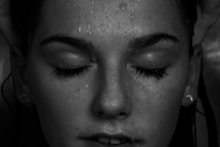 Schwarzweissfoto, Nahaufnahme Gesicht einer jungen Frau mit geschlossenen Augen und Wasser, das ihr von der Stirn runterläuft. Stichwort Taufe