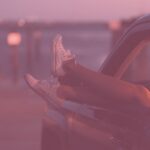 Füsse in weissen Turnschuhen werden aus einem Autofenster gestreckt, Abenddämmerung, im Hintergrund das Meer.