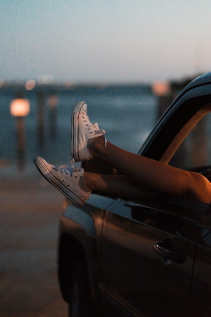 Autofenster im Abendlicht, woraus eine Person ihre Beine streckt, Füsse in weissen Stoff-Turnschuhen.