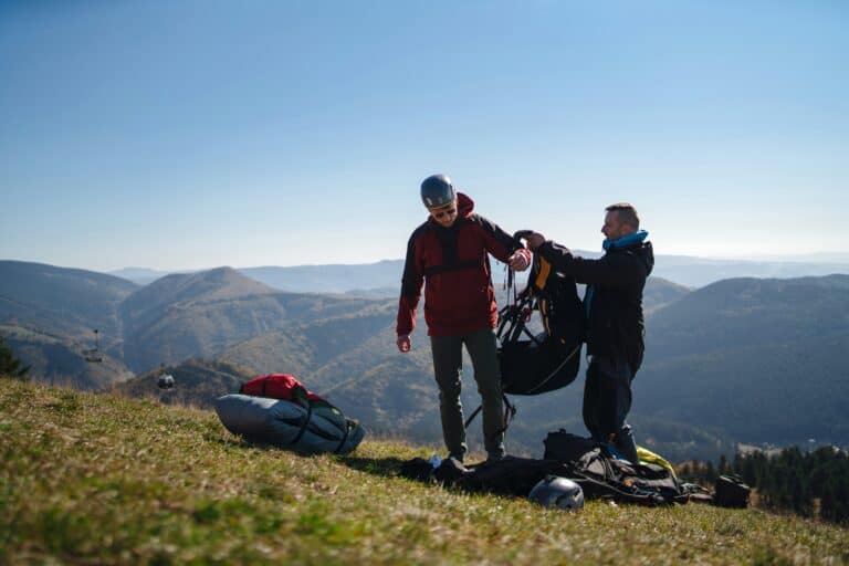 Zwei Männer auf einer Wiese vor Bergpanorama, der eine zieht dem anderen einen Gleitschirm-Rucksack an.