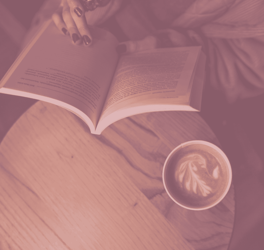 Blick von oben auf ein Buch, das von jemandem gelesen wird, daneben eine Tasse Cappuccino.