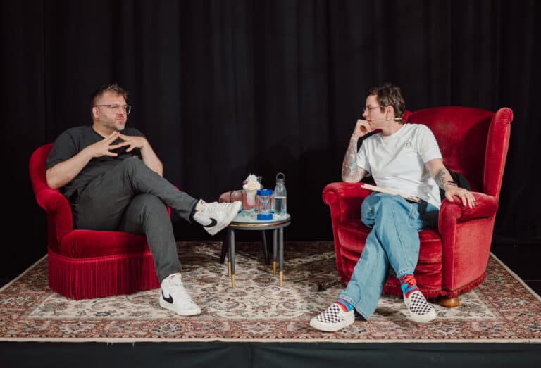 Ein Mann und eine Frau sitzen auf roten Sesseln vor einem schwarzen Vorhang