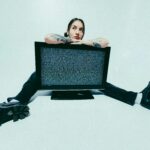 Studioaufnahme: Junge Frau vor weissem Hintergrund sitzt am Boden, zwischen ihren Beinen ein TV mit Bildstörung.