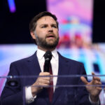 JD Vance, weisser Mann mit schwarzem Anzug, hält ernst dreinblickend Vortrag